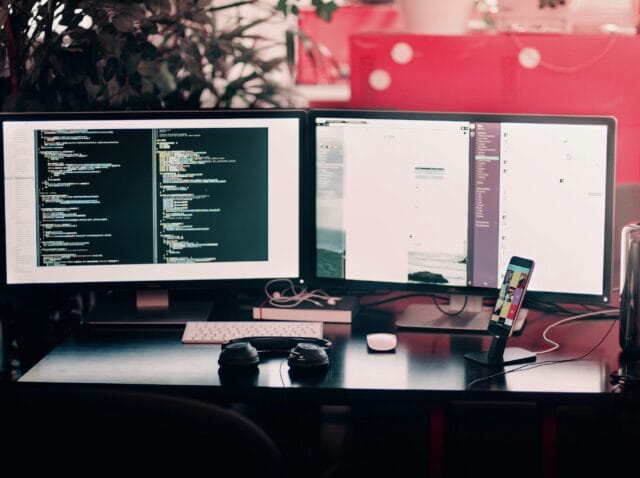Deux écrans d’ordinateur affichant du code et une interface, sur un bureau avec clavier, souris et smartphone en charge. Création site e-commerce chalon-en-champagne.